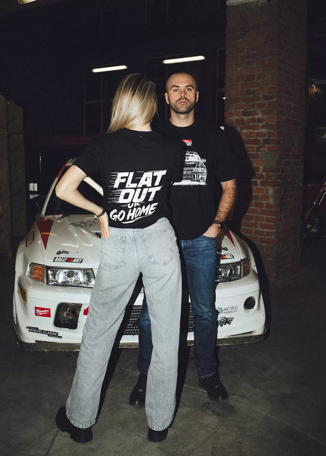Man and woman wearing black Flat Out or Go Home t-shirts in front of a white rally car