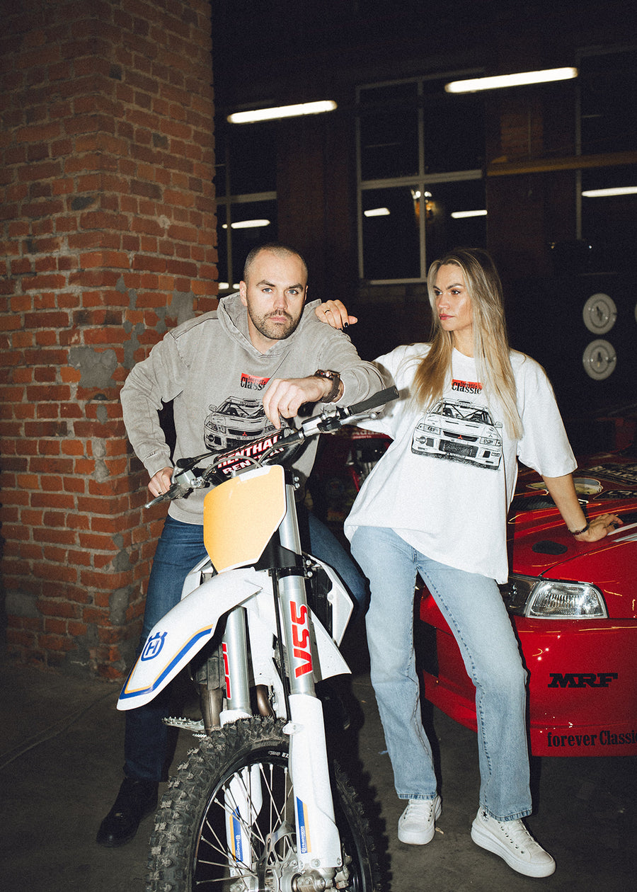 Couple posing with a Husqvarna motocross bike and the red rally car in a brick garage