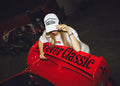 Model in a white Forever Classic cap and t-shirt leaning over the red rally car rear wing